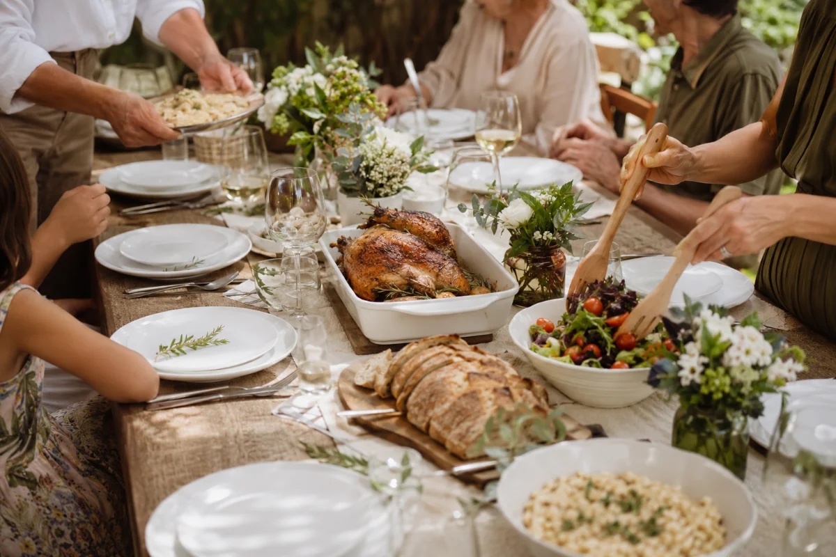 Fotografia lifestyle realista de um almoço de família em uma varanda/jardim com luz natural de meio-dia suave, mesa longa de madeira com toalha neutra, louças brancas, talheres, taças, arranjos discretos de flores e folhas, travessas com comida caseira elegante (salada colorida, frango assado, risoto, pão artesanal), pessoas de diferentes idades servindo e conversando com gestos naturais (sem rostos em destaque), clima acolhedor e sofisticado, composição equilibrada com profundidade de campo leve, foco na mesa e nas mãos, cores quentes e naturais, enquadramento horizontal 600x400, sem texto, sem logotipos, sem marcas d’água