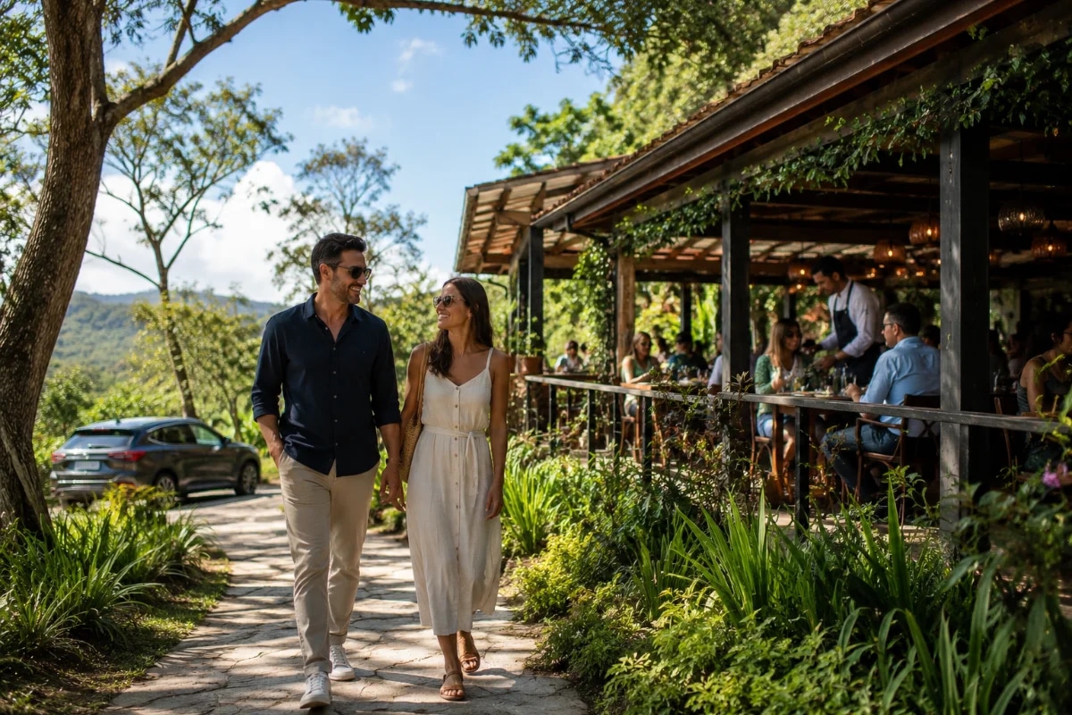 **Fotografia editorial realista de um casal de aproximadamente 30 a 45 anos chegando de carro a um restaurante sofisticado em meio à natureza na Serra da Cantareira durante o dia. O carro está estacionado discretamente ao fundo enquanto o casal caminha sorrindo por um caminho de pedra cercado por árvores e jardins bem cuidados em direção ao restaurante. Ao fundo, um ambiente elegante com varanda, mesas externas ocupadas por pessoas almoçando tranquilamente, garçons atendendo e vegetação exuberante criando sensação de refúgio. Céu azul com luz natural suave do fim da manhã, atmosfera de passeio de fim de semana e turismo de proximidade, transmitindo a ideia de "bate e volta gastronômico" e descanso sem necessidade de viagem longa. Composição horizontal 600x400, enquadramento cinematográfico, regra dos terços, foco nítido no casal em primeiro plano, profundidade de campo suave, cores naturais, estilo fotografia editorial premium, sem texto, sem logotipos, sem marcas d'água.**